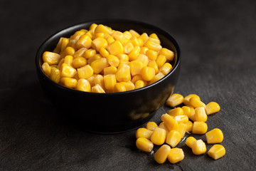 Canned sweet corn in a black ceramic bowl isolated on black slate next to spilled sweet corn.