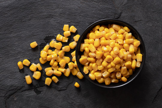 Canned sweet corn in a black ceramic bowl isolated on black slate next to spilled sweet corn. Top view.