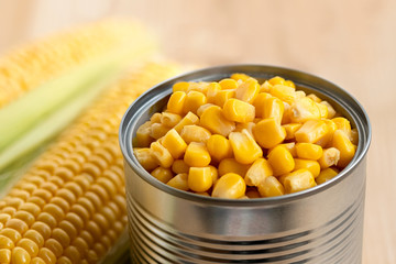 A can of sweet corn next to corn cobs on light wood.