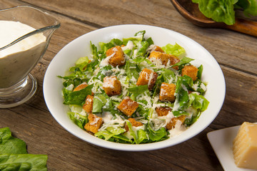 Caesar salad in a white bowl on an old rustic wooden table with leaves with a sauce boat and parmesan. Selected focus.
