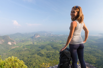 Young woman hiker stands on top of the mountain with backpack turns towards the camera and smiles