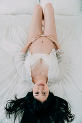 Naklejka premium upside down photo of beauty sexy pregnant asian japanese woman in lingerie and white lacy gown lying in cozy bed in bright room. beautiful future mom model smiling face camera with bare belly.