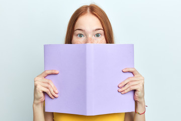 red-headed scared woman closing her face with a copy book, looking at the camera. close up portrait, isolated light blue background, education - 296077924