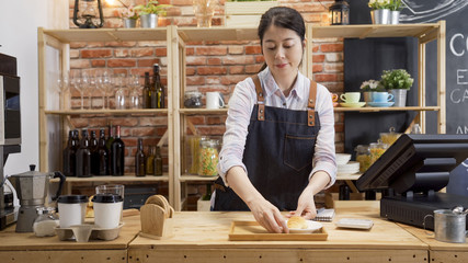 female bakery manager smiling while working at display counter. young girl waitress in cafe bar putting tasty handmade croissant on tray while preparing guest order. woman barista in coffee shop.