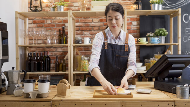 female bakery manager smiling while working at display counter. young girl waitress in cafe bar putting tasty handmade croissant on tray while preparing guest order. woman barista in coffee shop.