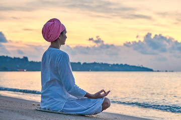 Woman in white clothes and a turban sits on a sandy beach at dawn and practices yoga