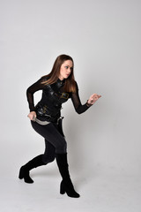 full length portrait of a pretty brunette woman wearing black leather fantasy costume. Standing pose, holding a dagger on a studio background.
