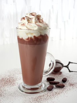 Hot chocolate latte with whipped cream, in tall glass, sprinkled with cocoa powder, on white background