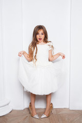 Little princess excited girl in fashion white dress wearing big mothers sparkle heels shoes on white background. Free space for text mockup