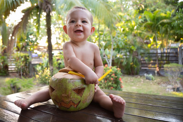 Fototapeta premium A small infant baby takes out a straw from a green coconut which is made in the shape of a halloween symbol.