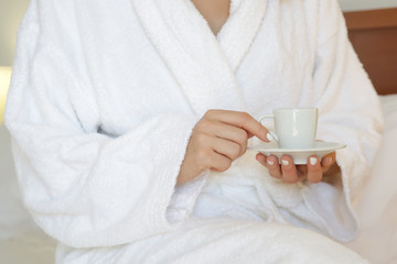 Woman in a white bathrobe having a cup of coffee in the bedroom