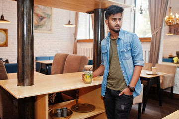 Portrait of handsome successful bearded south asian, young indian freelancer in blue jeans shirt standing in cafe with cocktail.