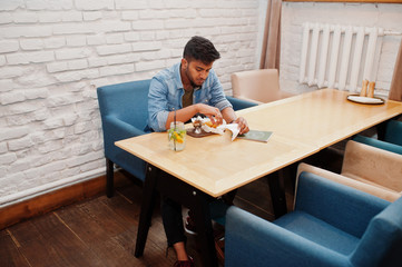 Portrait of handsome successful bearded south asian, young indian freelancer in blue jeans shirt sitting in cafe with chicken nuggets and lemonade.