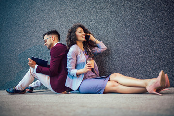 Young entrepreneurs sitting down in front of a modern office building and using phone. Brainstorming outside in a beautiful day.