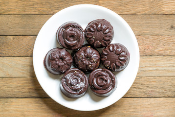 Delicious raw vegan sweets. Homemade chocolate dessert. Top view of natural harmless candies, sprinkled with coconut flakes, on white saucer on wooden table. Dietary tea treat. Copy space.