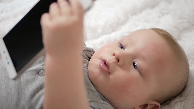 Cute Baby Holds Phone In Hands. The Child Plays With Cell Phone, Examines It, Tugs In His Hands And Tries To Eat Smartphone Putting It In His Mouth