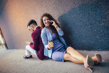 Young entrepreneurs sitting down in front of a modern office building and using phone. Brainstorming outside in a beautiful day.