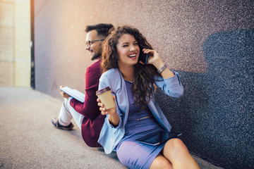 Young entrepreneurs sitting down in front of a modern office building and using phone. Brainstorming outside in a beautiful day.