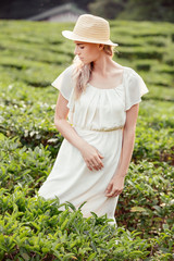 Portrait of a beautiful and romantic young blonde girl in white summer dress and hat posing while walking among green bushes in park during vacation on warm summer day. Concept of love for the natural