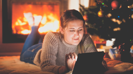 Toned portrait of smiling woman with tablet lying on floor next to firepalce and Christmas tree