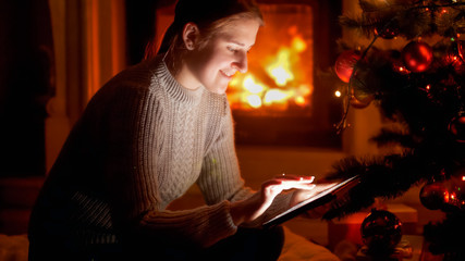 Portrait of smiling young woman sitting next to Christmas tree and typing message on tablet computer