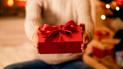 Closeup photo of young owman sitting on floor next to Christmas tree and showing red gift box in...