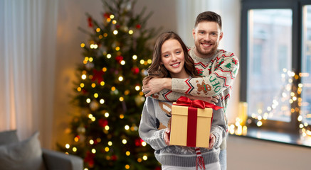 Obraz na płótnie Canvas people and holidays concept - portrait of happy couple with christmas gift at ugly sweater party over home background