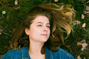 A beautiful young blonde girl with blue eyes with a blue jeans jacket lies on a green autumn moss...