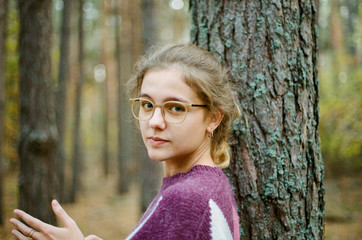 A young adult girl with blond hair gathered in a ponytail, wearing glasses stands in the autumn forest against a background of trees, holds her hands in front of herself, looks directly at the camera