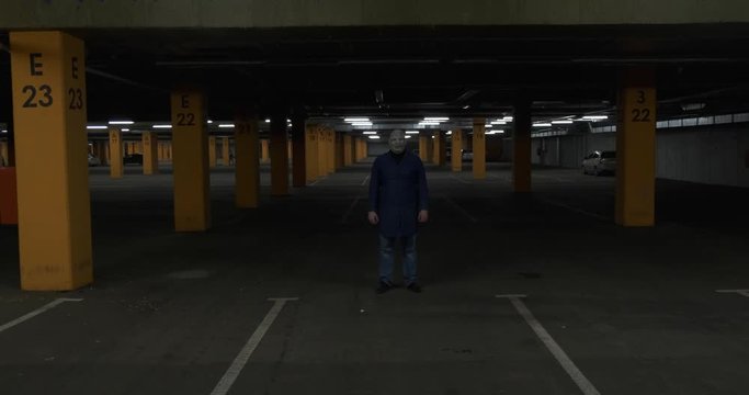 Camera on cart slowly drives up to face of adult man. He is wearing mask with terrible scars and crooked teeth, lonely figure in parking lot, darkness around, depressing atmosphere, dangerous maniac