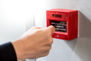 Male hand punching red fire alarm switch on concrete wall in public building. Industrial fire warning system equipment for emergency.