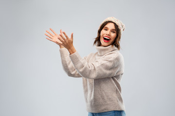 winter holidays and people concept - happy smiling young woman in hat and sweater with raised hands holding something imaginary over grey background