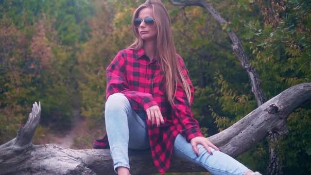 Slow motion of a sexy girl taking off glasses outside in nature, passionately looking at camera, sitting on a log. Forests and tourism concept.