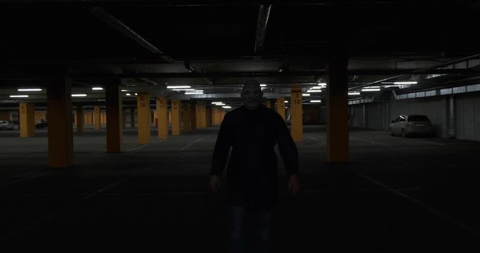 Camera on cart slowly moves away from man in scary halloween costume of movie killer walking towards her, maniac slowly approaches viewer, waddles around, stops in beam of light, leather mask scares