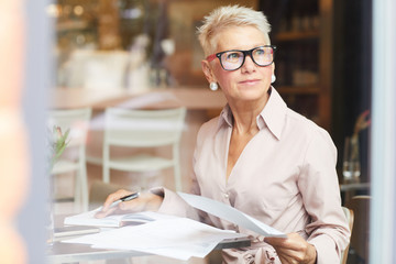 Mature businesswoman with blond short hair in eyeglasses sitting at the table and working with...
