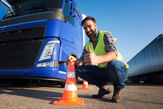 Shot Of Bearded Man Learning How To Drive Truck At Driving Schools. Truck Driver Candidate Training For Driving License. Standing By The Traffic Cone In Reflective Vest. Truck In The Background.