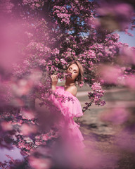 Art fashion portrait of a beautiful young blonde woman in a spring pink blooming garden in a long...