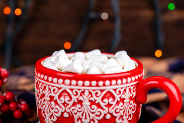 Close up of red mug with ornament with hot cocoa drink and white sweet marshmallows with garland bokeh lights background. Christmas and New Year holidays concept.