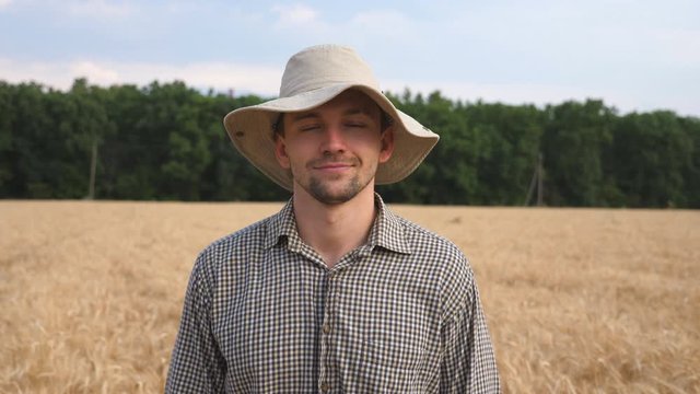 Close up of happy smiling farmer in hat looking into camera against the blurred background of wheat field. Portrait of handsome young agronomist standing in the barley meadow. Agriculture concept