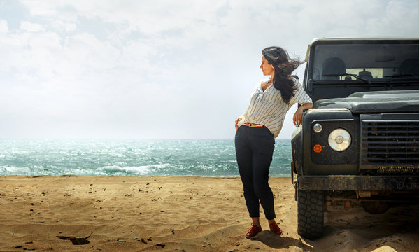 Attractive Young Woman Traveler Enjoying The Sea View, Leaning Back On a Classic Car SUV. Adventure Travel Concept and Lifestyle with copy-space