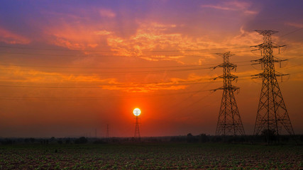Obraz premium high voltage post and high voltage tower electric transmission tower system with blue sky sunset silhouette with blue sky sunset, white clouds and meadow and grass in the evening.