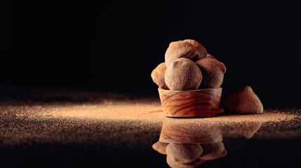 Chocolate truffles in small wooden dish sprinkled with cocoa powder. Black reflective background.