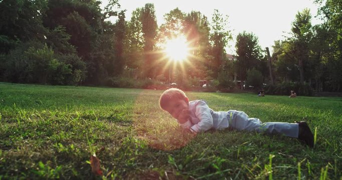 Charismatic small boy two years old falling down on the green grass and looking straight to the camera smiling very cute