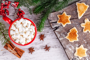 Cozy winter breakfast concept. Red cup with hot chocolate and marshmallows, cinnamon sticks, anise stars, toasted shaped bread slices on wooden table in Christmas decorations. New Year morning food.