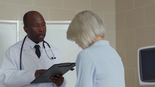 African American doctor in white coat holding clipboard with lab test results, telling young female patient about scary diagnosis and trying to comfort her