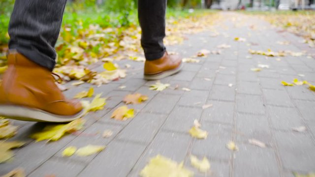 A young man in leather shoes is walking along a sidewalk with fallen leaves. Fall season. Outdoor city walk concept. Slow motion
