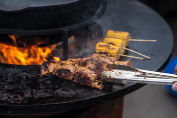 cooking, frying meat and corn in a huge flat pan with an open fire in the middle