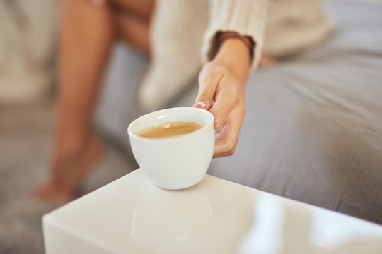 Cropped image of caucasian woman in beige sweater putting cup of coffee on night  table. bedroom interior. Selective focus on cup.