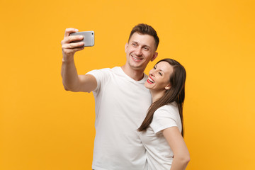 Smiling young couple friends guy girl in white empty blank design t-shirts posing isolated on yellow orange background. People lifestyle concept. Mock up copy space. Doing selfie shot on mobile phone.
