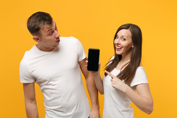 Young couple two friends guy girl in white t-shirts posing isolated on yellow orange background. People lifestyle concept. Mock up copy space. Pointing finger on mobile phone with blank empty screen.